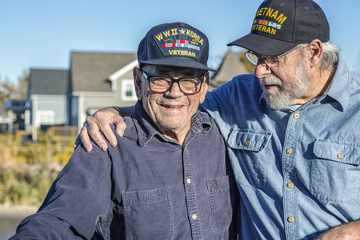Two elder men wearing "Veteran" hats, smiling outdoors, one has an arm around the other.